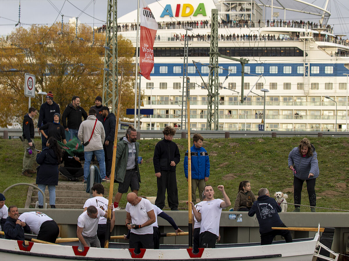 im Vordergrund steigen änner in einen Kutter. Dahinter am Wiesenhang stehen Menschen. Dahinter liegt ein AIDA-Kreuzfahrtschiff. An jedem Deck stehen Menschen wie an einer Perlenschnur aufgereiht.