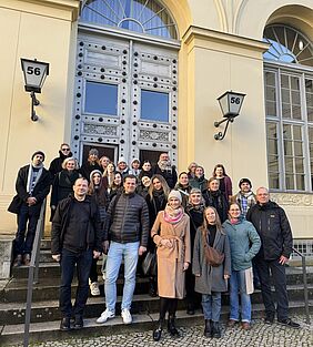 Participants at the T.A.B. conference in Berlin. About 30 people are standing in several rows on the steps in front of a historic building with a tall double door and large windows.