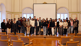 Around 35 people stand in a bright hall in front of a projection screen. Mayor Beyer stands in the middle of the front row, with Prof. Dr Arnaout on the far right.