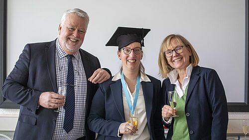 Alle drei PErsonen stehen nebeneinander, halten ein Sektglas (mit alkoholfreiem Sekt) in den Händen und strahlen freudig in die Kamera.