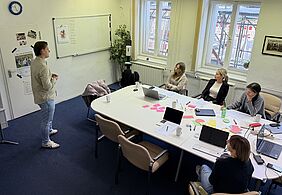Interdisciplinary teamwork in the Creative Sprint. Several people are seated at a conference table with laptops, notes and presentation cards, while one person stands at the front and gives a presentation.