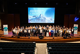 Around 100 international participants are posing for a group photo on a stage in front of a large presentation screen at the Aurora Symposium.