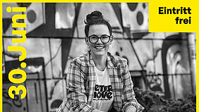 Eine Frau mit dunkler Brille lächelt ins Bild, sie trägt eine karierte Jacke
