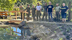 Gruppe von acht Personen steht auf einem Weg neben einem kleinen Holzsteg, der über einem Bach führt. Im Hintergrund sind Büsche zu sehen.