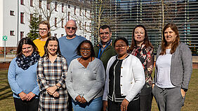 Die neun Personen stehen in zwei Reihen. Im Hntergrund ist die Hochschulbibliothek und ein Lehrgebäude zu sehen, das weiß in der Sonne leuchtet.