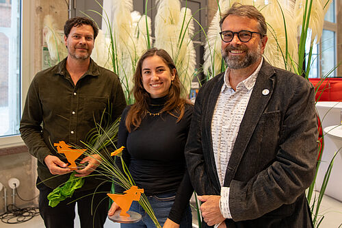Drei Personen stehen nebeneinander in einem Raum vor Gräsern. Zwei halten orangefarbene Fischer-Figuren als Auszeichnungen und Sträuße in der Hand. Alle lächeln.