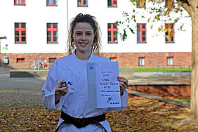 Die Studentin ist in Karatekleidung mit ihrer Urkunde und der Bronzenadel in der Hand vor dem Haus 1 zu sehen, wo viele ihrer Lehrveranstaltungen stattfinden.