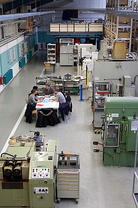 Man schaut von oben in eine grße Halle. Es sind mehrere MAschinen zu sehen. An einem Tisch sitzen 5 Personen, die auf die ausgebreiteten Zeichnungen und Tabellen schauen.