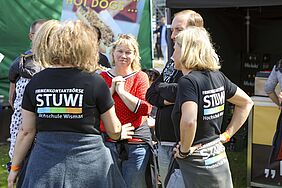 Zwei Frauen sind von hinten zu sehen. Sie haben T-Shirts mit dem StuWi-Logo an und unterhalten sich mit einer Besucherin.