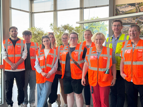 Group photo of students wearing orange high-visibility vests in the entrance hall. The individuals are standing in a line. Mr Mertens, the contact person for the company visit, is second from the right.
