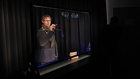 Im dunklen Raum ist leuchtender Text auf einer Glasscheibe zu sehen, hinter der eine Person steht.