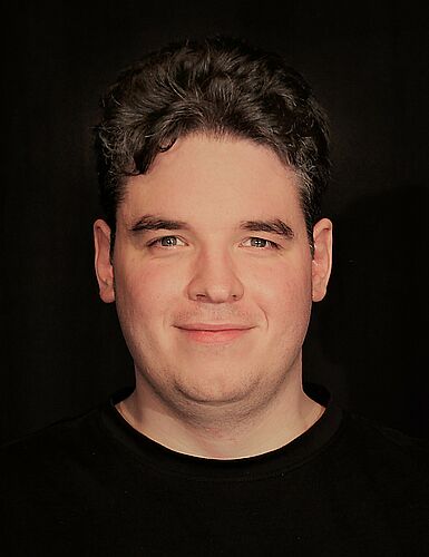 Porträtfoto eines lächelnden jungen Mannes mit dunklem Haar, dunklem Pullover und schwarzem Hintergrund