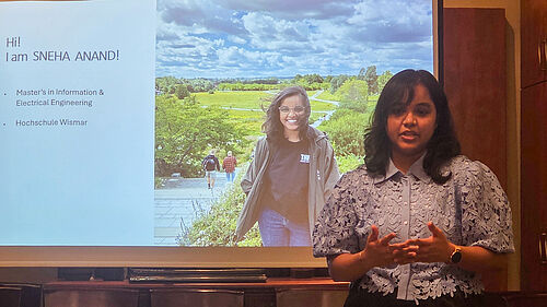 Eine Frau schaut in die Kamera, wobei sie mit ihren Händen gestikuliert. Hinter ihr ist eine Leinwand mit einer Präsentation zu sehen. Auf dieser steht ihr Name sowie was und wo sie studiert.
