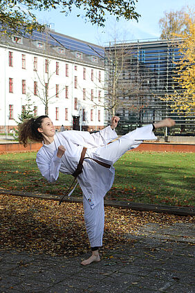 Es ist eine Sportlerin in Karatekleindungauf dem Campus zu sehen. Sie zeigt einen Karate-Seitwärtskick, so dass ein Fu über ihren Kopf reicht.