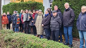 Auf dem Gehweg zwischen geschnittenen Hecken steht eine Gruppe von Personen aufgereiht und schaut in die Kamera.