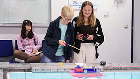 Ein Schüler (links im  Bild) hebt einen Modellcontainer mittels einer magnetischen Angel auf sein DIY-Schiff. Seine Teamkameradin steht daneben und hält die Fersteuerung in der Hand.