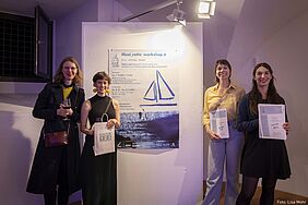 Die drei Preisträgerinnen stehen vor einem Ausstellungsbanner und halten ihre Urkunden in die Kamera. Mit ihenen auf dem Bild ist auch Professorin Martineck zu sehen.