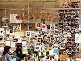 vor einer Atelierwand, an der sich zahlreiche Fotos und Skizzen befinden, sitzen zwei Studentinnen.