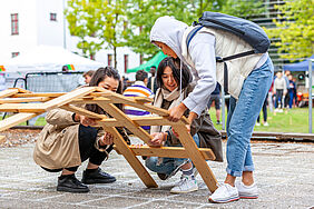 Drei Studentinnen versuchen gemeinsam Holzlatten zu einer Leonardo-Brücke zusammenzufügen.