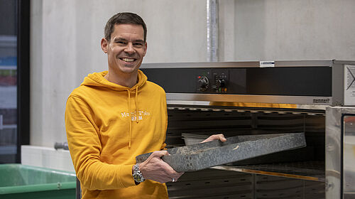 Der Professor steht vor einem Schrank, der wie ein großer Ofen aussieht. Er hält ein Blech in der Hand, das einem Backblech ähnlich sieht, und schiebt es in den Schrank.