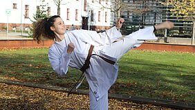 Es ist eine Sportlerin in Karatekleindungauf dem Campus zu sehen. Sie zeigt einen Karate-Seitwärtskick, so dass ein Fu über ihren  Kopf reicht.