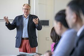 Der REktor gestikuliert mit ausgestreckten Händen. Am rechten Rand sieht man unscharf einge TEilnehmende, die in seine Richtung blicken.