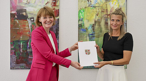 Die beiden Frauen stehen vor einer Wand mit zwei großen Bildern. Sie halten gemeinsam die Ernennungsurkunde in der Hand.