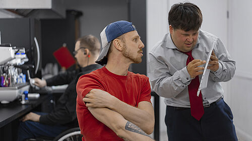 Es ist ein junger Mann im Rollstuhl zu sehen, der mit einem anderen jungen Mann spricht, der eine A4-Folie in der Hand hält.