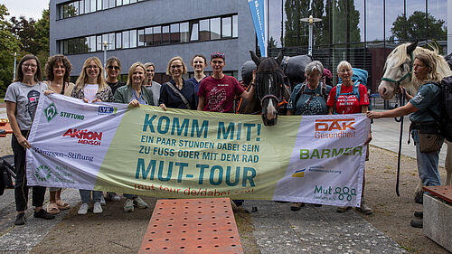 Zwei Pferde und mehrere Menschen stehen nebeneinander. Vor sich haben sie ein Banner ausgebreitet, auf dem die Aufforderung zur Teilnahme an der Mut-Tour steht: "Komm mit!" Außerdem sind die Logos der Unterstützereinrichtungen zu sehen. 