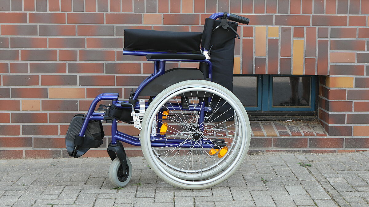 Der Rollstuhl steht vor einer Hauswand mit roten Klinkern. 