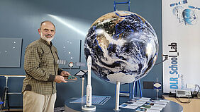 Albrecht Weidemann hält einen Modellsatelliten in der Hand und schaut dabei in die Kamera. Neben ihm steht ein Globus mit rund einem Meter Durchmesser.