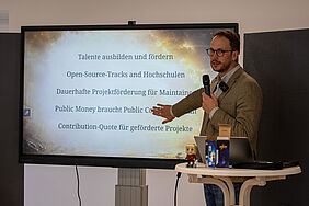 Die PErson steht mit einem Mikrofon in der Hand vor einem Bildschirm und zeigt mit der anderen Hand auf eine Textzeile.