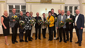 Die 13 Personen stehen nebeneinander. Die Ausgezeichneten halten Blumensträuße und Urkunden in der Hand.