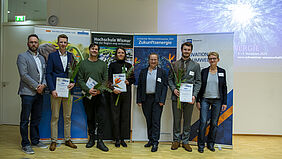 Alle Personen stehen nebeneinander. Die Preisträger halten Urkunden und Blumen in den Händen. Im Hintergrund sind vier Banner zu sehen.