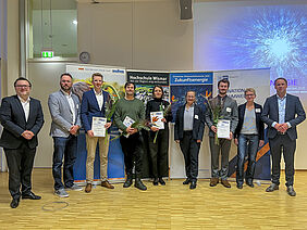 Alle Personen stehen nebeneinander. Die Preisträger halten Urkunden und Blumen in den Händen. Im Hintergrund sind vier Banner zu sehen.