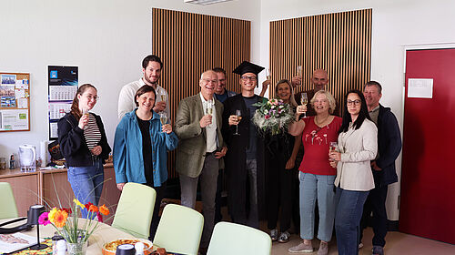 Dominic Strube steht nach seiner erfolgreichen Promotion im Kreis von Kolleginnen und Kollegen. Die Gruppe hält Sektgläser in den Händen und feiert den Abschluss der Dissertation. Dominic Strube trägt einen Doktorhut und hält einen Blumenstrauß.