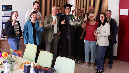 Dominic Strube steht nach seiner erfolgreichen Promotion im Kreis von Kolleginnen und Kollegen. Die Gruppe hält Sektgläser in den Händen und feiert den Abschluss der Dissertation. Dominic Strube trägt einen Doktorhut und hält einen Blumenstrauß.
