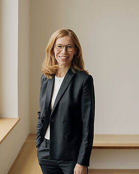 Die Frau steht in der Ecke eines Raumes und lächelt in die Kamera.