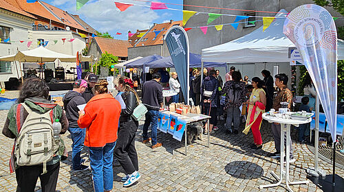 Es ist ein weißer offener Zeltpavillon zu sehen mit einem Stehtisch davor. Daneben befinden sich weitere Pavillons und im Hintergrund eine Bühne. Etliche Menschen stehen und laufen im Hof. Über ihnen when bunte Fähnchen an Stricken.