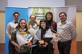 Sechs Personen stehen vor zwei Bannern mit Text, zwei Frauen halten Blumensträuße und Urkunden, alle lächeln in die Kamera.
