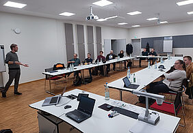 Ralmo Bode stands at the front of the seminar room and presents his research work. A slide with diagrams can be seen in the background, and the participants listen attentively.