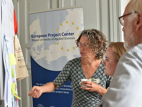 Three people are standing in front of a pinboard with notes, discussing its content. A woman with curly hair and glasses is explaining something and pointing at the board while holding a cup. In the background, there is a roll-up banner reading “European Project Center – Wismar University of Applied Sciences” with a map of Europe and yellow stars. The scene appears to be an engaged discussion during a workshop or project meeting.