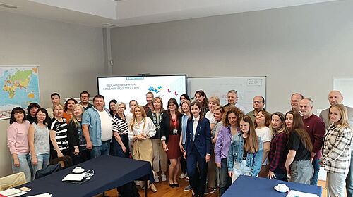 A group of around 40 participants from various countries stand together in a seminar room in front of a screen showing the EUComplianceM4UA project title.