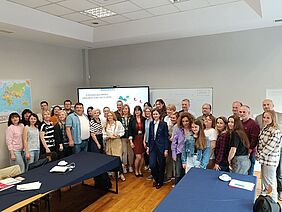 A group of around 40 participants from various countries stand together in a seminar room in front of a screen showing the EUComplianceM4UA project title.