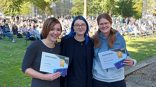 Es ist die AStA-Vorsitzende, Anna Dahlke, mit den beiden Preisträgerinnen Julia Heuer und Martina Schindler zu sehen. 