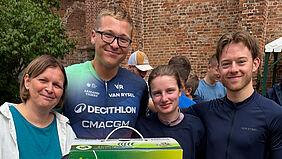 Eine Gruppe von vier Personen steht vor einer Backsteinmauer. Die zwei Frauen und zwei Männer lächelnd in die Kamera. Einer trägt erkennbar ein Fahrradtrikot