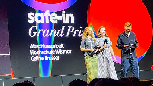 Über Köpfe hinweg schaut man auf eine Bühne. Drei Personen stehen auf einer Bühne vor einer großen Leinwand mit der Aufschrift "safe-in Grand Prix Abschlussarbeit Hochschule Wismar Celine Brussel"..