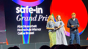 Über Köpfe hinweg schaut man auf eine Bühne. Drei Personen stehen auf einer Bühne vor einer großen Leinwand mit der Aufschrift "safe-in Grand Prix Abschlussarbeit Hochschule Wismar Celine Brussel"..