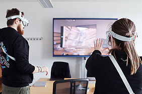 Two students wearing Apple Vision Pro headsets are standing in a seminar room. One of them interacts with a virtual scene displayed on a large screen by raising both hands.