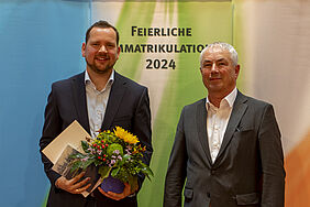Der Preisträger hält Blumen und Urkunde in der Hand. Beide Personen stehen vor einem farbigen Banner, auf dem in der Mitte steht:"Feierliche Immatrikulation 2024".